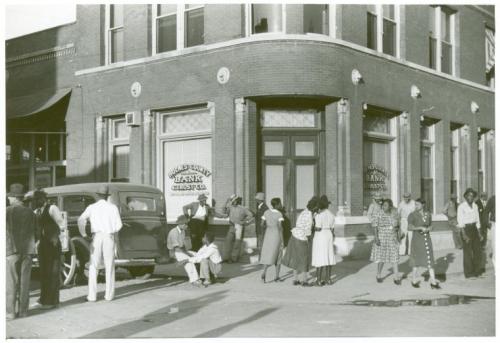 Historic black-and-white photo of a bank with people gathered outside, some seated and others standing, in a city setting.