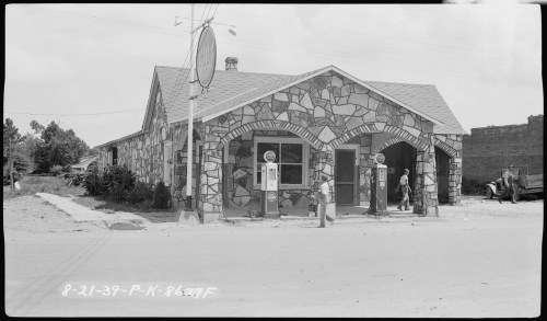 Historic stone building with gas pumps outside, two people nearby, and a vintage car parked on the street.