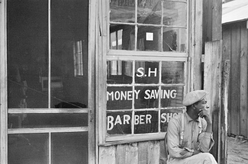 A man sits pensively outside a barber shop with a sign reading "S.H. Money Saving Barber Shop." Black and white photo.