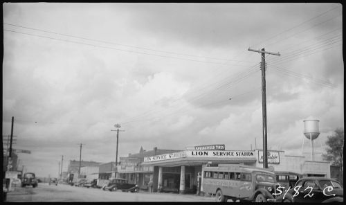Black and white photo of a street scene featuring the Lion Service Station and vintage cars under a cloudy sky.