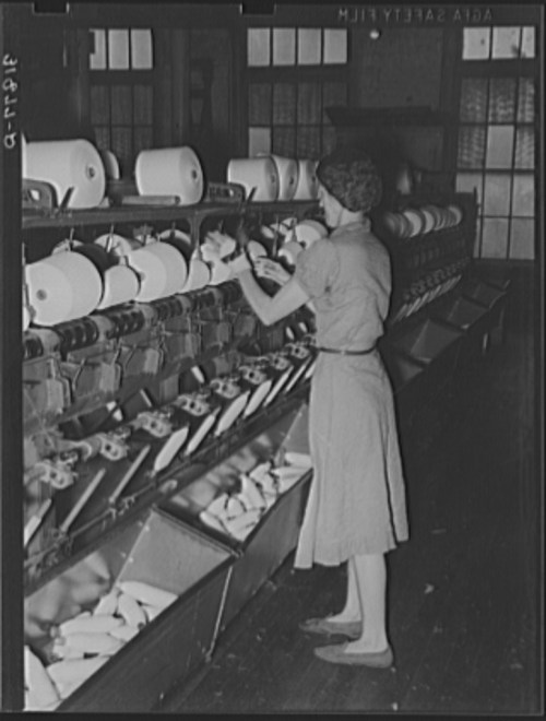 A woman operates machinery in a textile factory, handling spools of thread in a historical black-and-white photo.