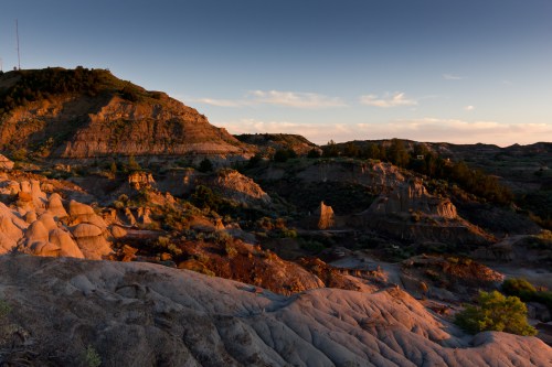 Sunset over rugged hills and rock formations, with warm light illuminating the landscape and scattered vegetation.