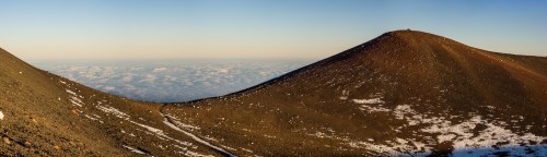 Panoramic view of a volcanic landscape with a smooth, sloping cone and a clear sky above. Snow patches visible.