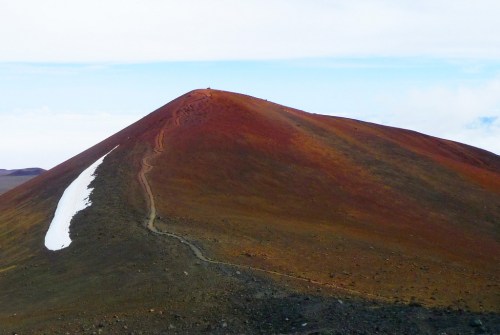 A colorful mountain peak with a winding path and a small patch of snow against a blue sky.