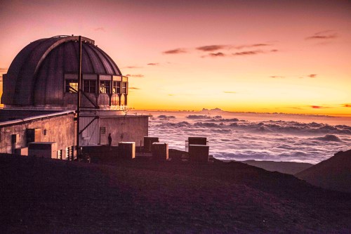 A dome-shaped observatory at sunset, overlooking a sea of clouds with vibrant orange and purple hues in the sky.