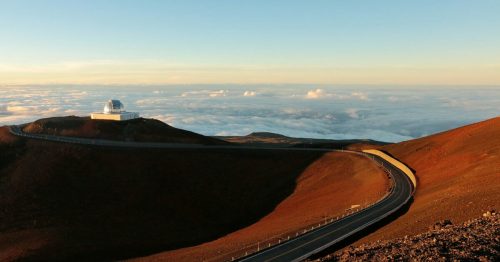 A winding road leads to a white observatory atop a mountain, surrounded by clouds and a colorful landscape at sunrise.