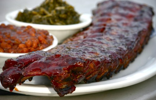 A plate featuring a glazed rib rack alongside bowls of baked beans and collard greens.