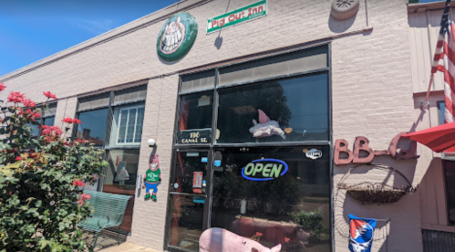 Exterior of a restaurant with a pig sign, large windows, and an "OPEN" sign, surrounded by flowers and flags.