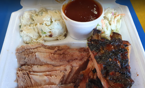 A takeout container with sliced beef, barbecue ribs, mashed potatoes, coleslaw, and a cup of barbecue sauce.