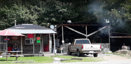 A rustic roadside building with a porch, a pickup truck parked outside, and smoke rising from a nearby structure.