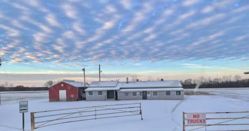 A snowy landscape featuring a red barn, a house, and a "No Trucks" sign under a cloudy sky at dawn.