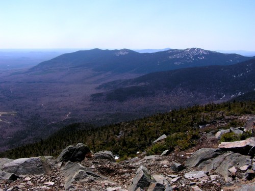 A panoramic view of mountains and valleys under a clear blue sky, with rocky terrain in the foreground.