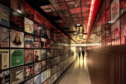 A narrow hallway lined with colorful album covers, illuminated by red lights, leading to a distant exit.