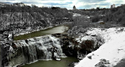 A snowy landscape featuring a waterfall cascading into a river, with a distant building and overcast sky.