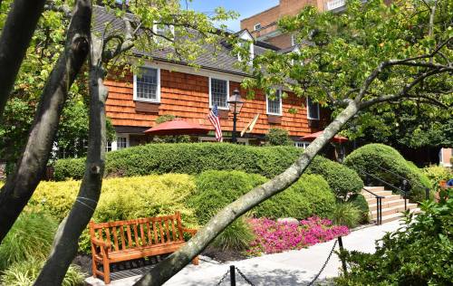 A charming house with shingle siding, surrounded by lush greenery and colorful flowers, featuring a wooden bench in the foreground.