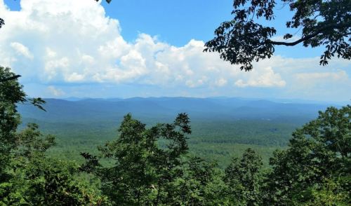 A panoramic view of lush green mountains under a bright blue sky with fluffy white clouds.