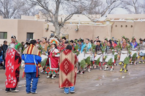 A vibrant procession of Indigenous dancers in traditional attire, celebrating with greenery and colorful decorations.