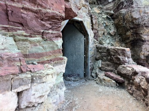An entrance to a cave or rock formation, surrounded by layered red and beige stone.