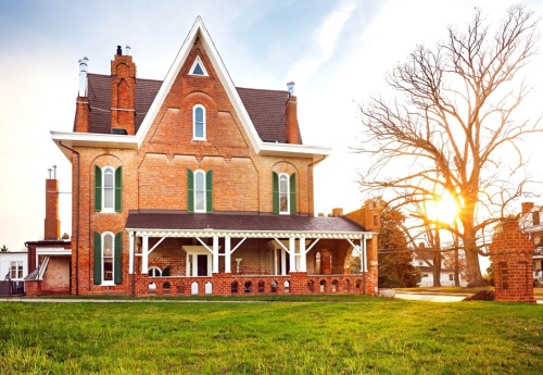A large, historic brick house with a pointed roof, surrounded by green grass and a tree, illuminated by a sunset.