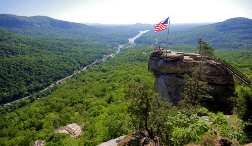 A scenic view from a rocky overlook featuring a large American flag, surrounded by lush green mountains and a winding river.
