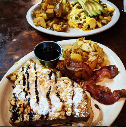 A plate of French toast topped with nuts and syrup, alongside crispy bacon and seasoned potatoes. Another plate of scrambled eggs with avocado.