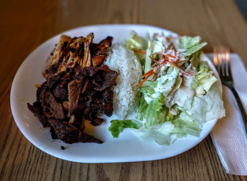 A plate of grilled chicken and beef served with white rice and a side salad drizzled with dressing.