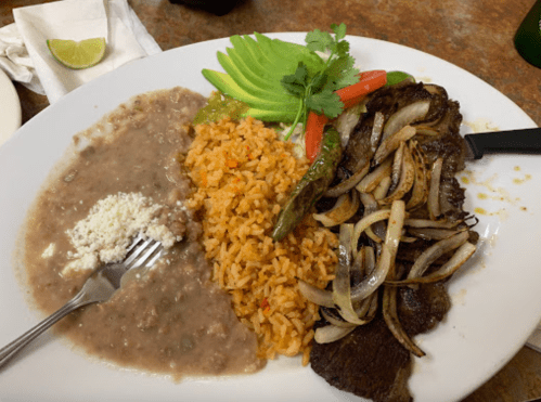 A plate of Mexican food featuring grilled steak, rice, refried beans, avocado, onions, and garnishes.