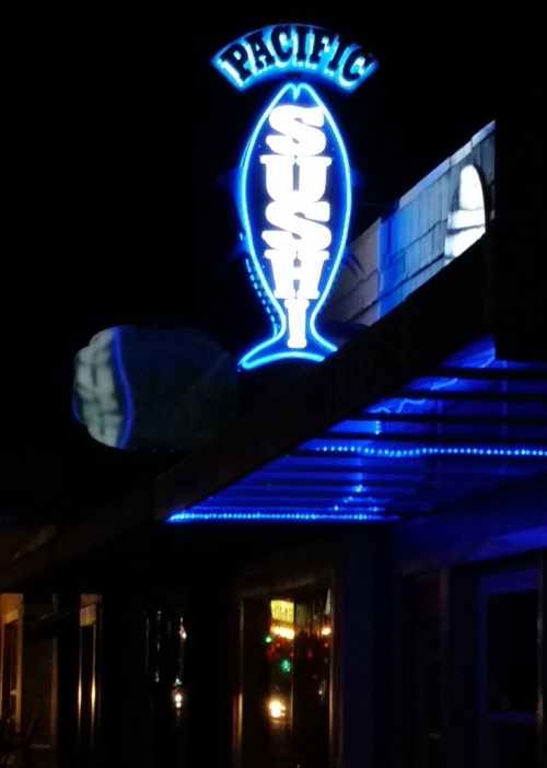 Neon sign for "Pacific Sushi" illuminated at night, featuring a fish design.