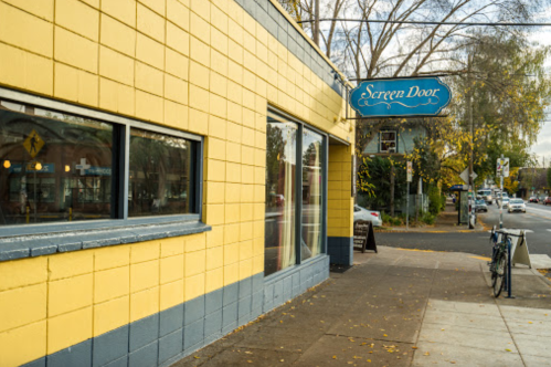 A bright yellow building with a sign reading "Screen Door," located on a street with trees and parked bicycles nearby.