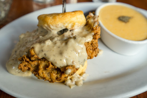 A plate featuring a biscuit topped with fried chicken and creamy gravy, alongside a small bowl of yellow grits.