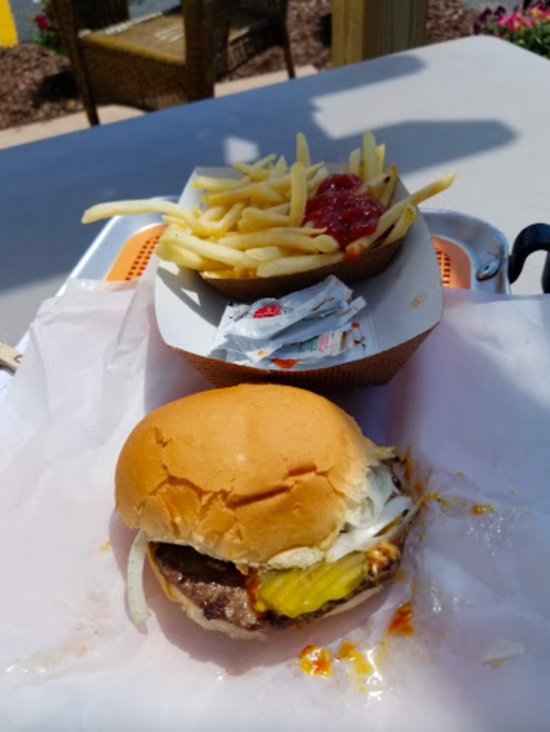 A close-up of a hamburger with pickles and onions, next to a container of fries and ketchup on a table.