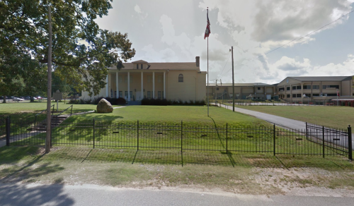 A large building with columns and a flag, surrounded by green grass and a fence, with another structure in the background.