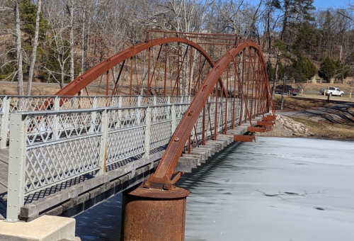 A rusted metal bridge arches over a frozen lake, surrounded by bare trees and a clear blue sky.