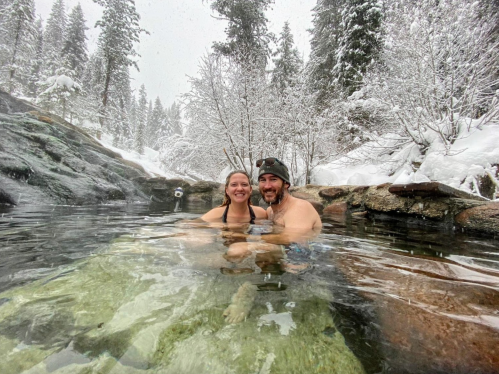 A couple smiles while relaxing in a hot spring surrounded by snowy trees and rocks.
