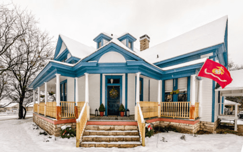A blue and white house with a snow-covered yard, featuring a Marine Corps flag and festive decorations.
