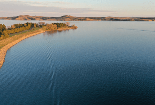 A serene lake at sunset, with gentle ripples and lush greenery along the shore, surrounded by rolling hills.