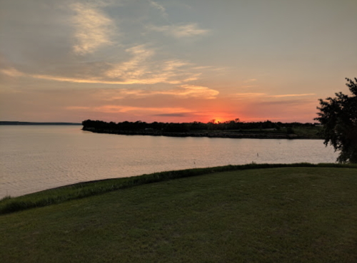 A serene sunset over a calm lake, with a grassy shoreline and trees silhouetted against the colorful sky.