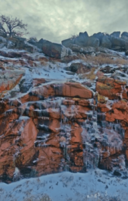 A rocky cliff with ice formations and patches of snow under a cloudy sky.