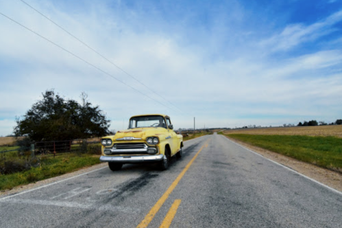 A vintage yellow truck parked on a rural road with fields and a cloudy sky in the background.