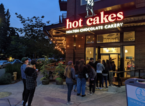 A line of people waiting outside Hot Cakes, a molten chocolate bakery, in the evening with warm lights glowing.