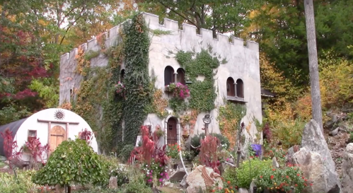 A whimsical stone building covered in ivy, surrounded by colorful flowers and trees in a vibrant autumn setting.