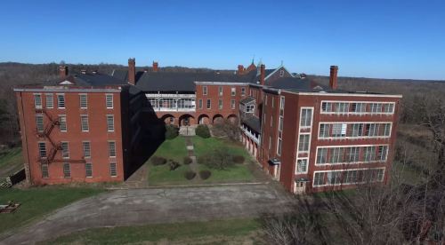 Aerial view of a large, abandoned brick building complex surrounded by greenery under a clear blue sky.