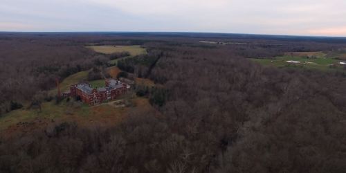 Aerial view of a large, abandoned brick building surrounded by dense trees and open fields.