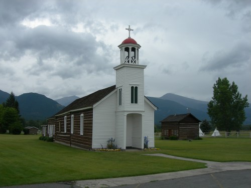 A small white church with a red roof and bell tower, surrounded by green grass and mountains under a cloudy sky.