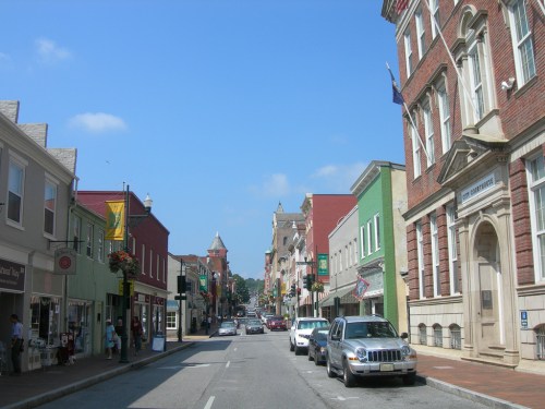 A charming street lined with colorful buildings, shops, and parked cars under a clear blue sky.