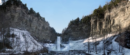 A panoramic view of a frozen waterfall surrounded by rocky cliffs and trees, with snow covering the ground.