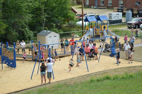 A busy playground filled with children and adults, featuring swings, slides, and a play structure surrounded by greenery.