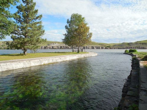 A serene river scene with trees, a grassy bank, and gentle ripples under a partly cloudy sky.