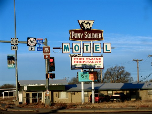 Sign for Pony Soldier Motel with "Vacancy" and "High Plains Hospitality" under a clear blue sky. Traffic lights visible.