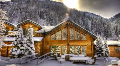 A cozy log cabin surrounded by snow-covered trees and mountains, with large windows reflecting the winter landscape.